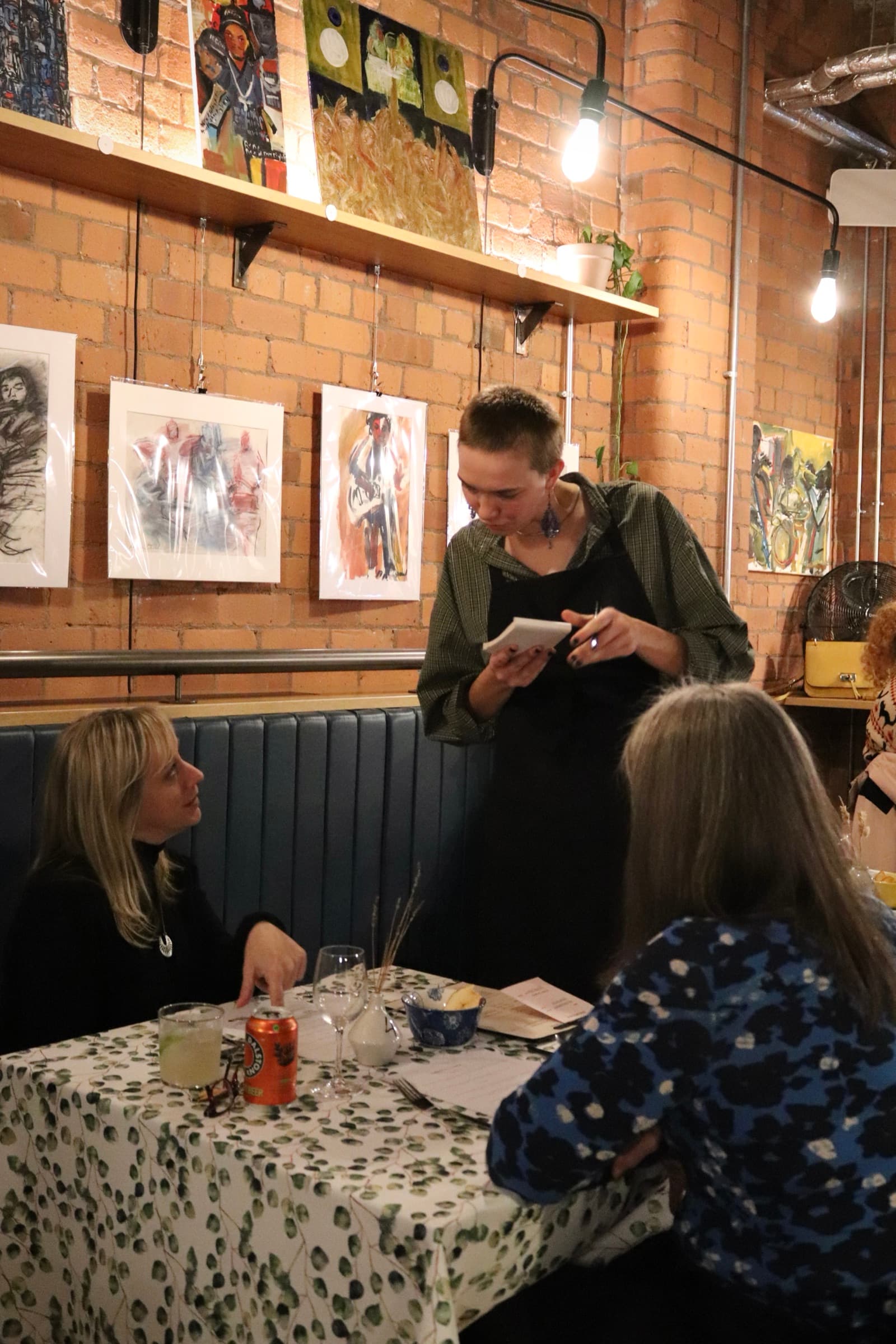 A server smiling as they take an order from two guests at a candle-lit table beneath a wall of paintings
